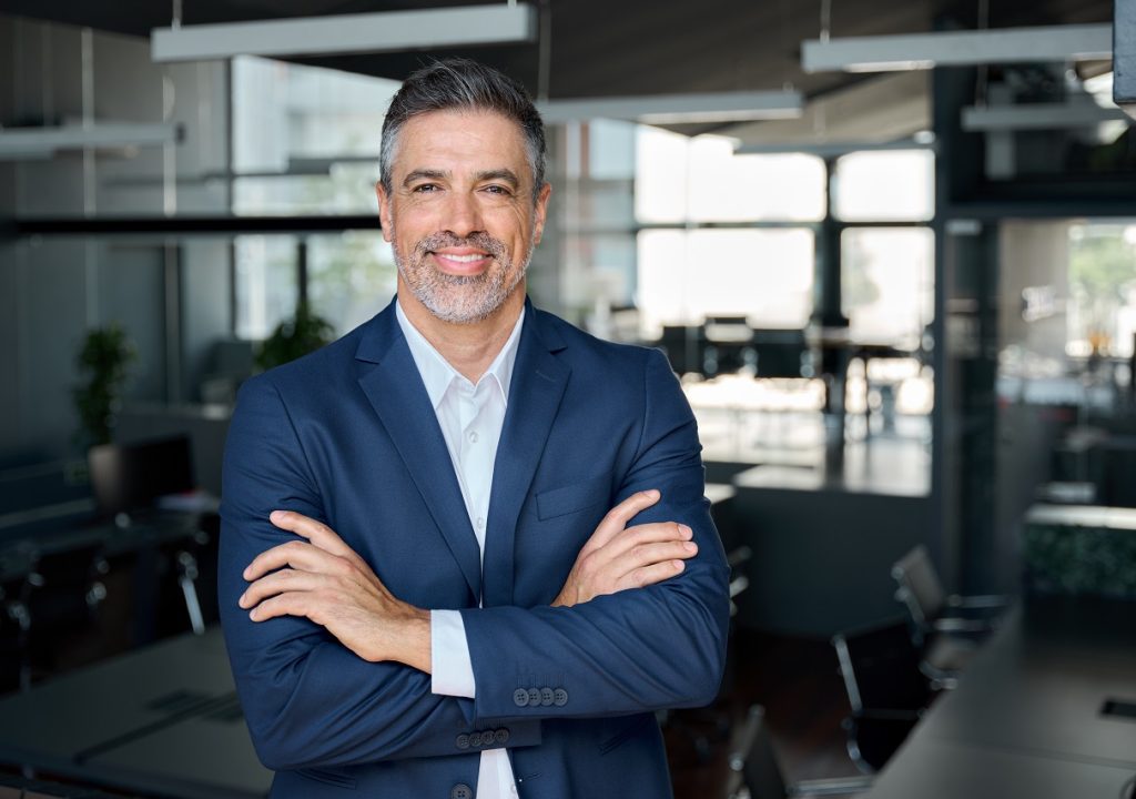 Happy middle aged business man ceo standing in office arms crossed. Smiling mature confident professional executive manager, proud lawyer, confident businessman leader wearing blue suit, portrait.